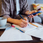 Person writing in a notebook while holding a smartphone, with bills and a cup of coffee on the table.