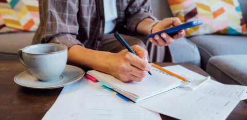 Person writing in a notebook while holding a smartphone, with bills and a cup of coffee on the table.