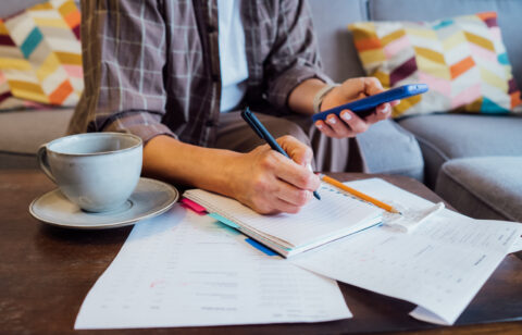 Person writing in a notebook while holding a smartphone, with bills and a cup of coffee on the table.