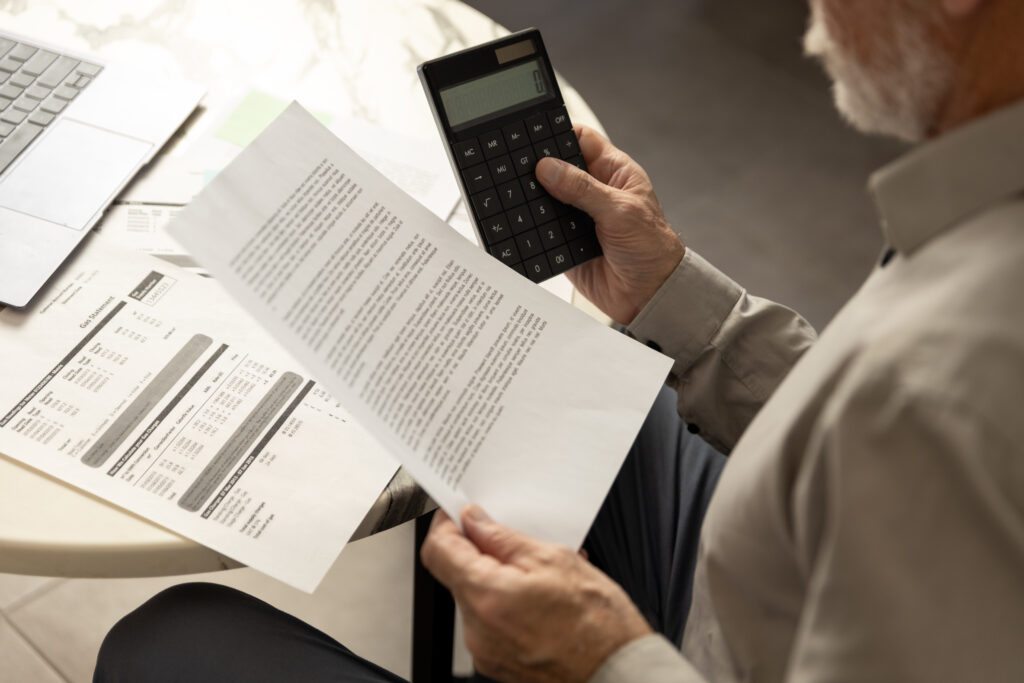 Older man reviewing a bill while holding a calculator at a table with a laptop nearby.