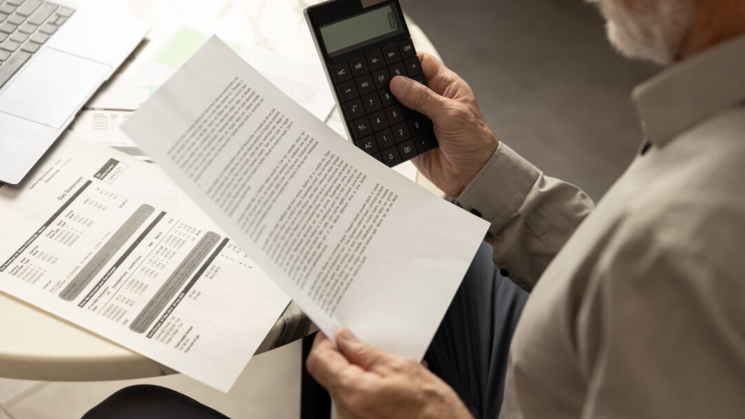 Older man reviewing a bill while holding a calculator at a table with a laptop nearby.