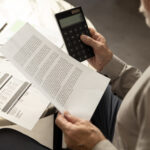 Older man reviewing a bill while holding a calculator at a table with a laptop nearby.