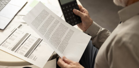 Older man reviewing a bill while holding a calculator at a table with a laptop nearby.