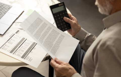 Older man reviewing a bill while holding a calculator at a table with a laptop nearby.