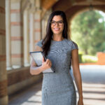 Woman holding a tablet and smiling while standing in a covered outdoor walkway.