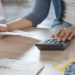 Person using a calculator while reviewing bills and holding a document at a desk.