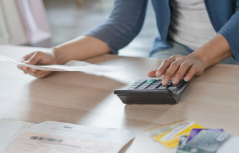Person using a calculator while reviewing bills and holding a document at a desk.