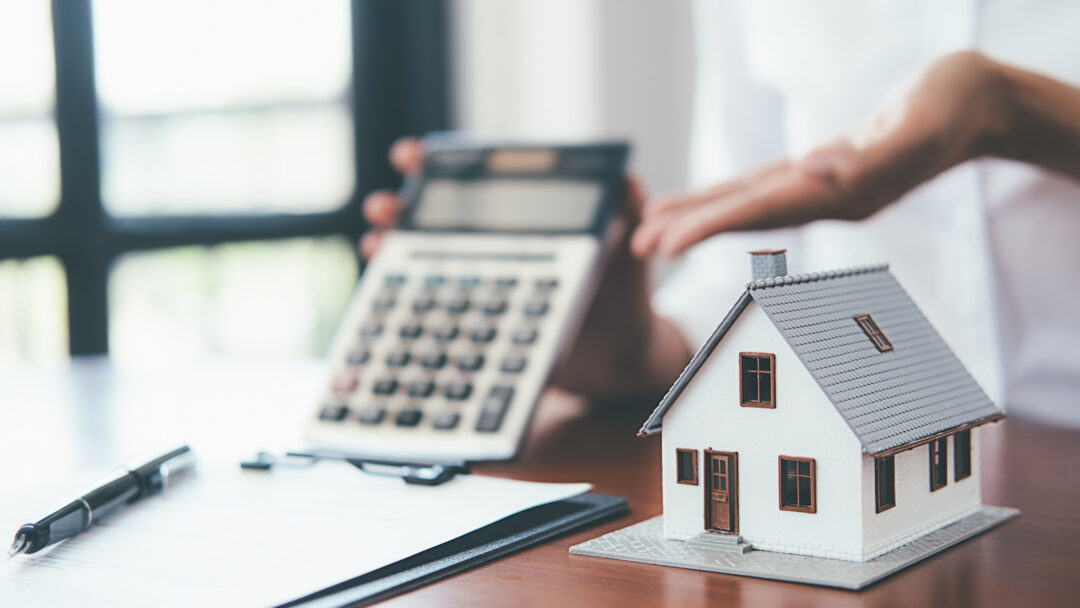 Small model house on a desk with a calculator and paperwork in the background.