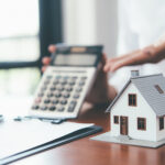 Small model house on a desk with a calculator and paperwork in the background.
