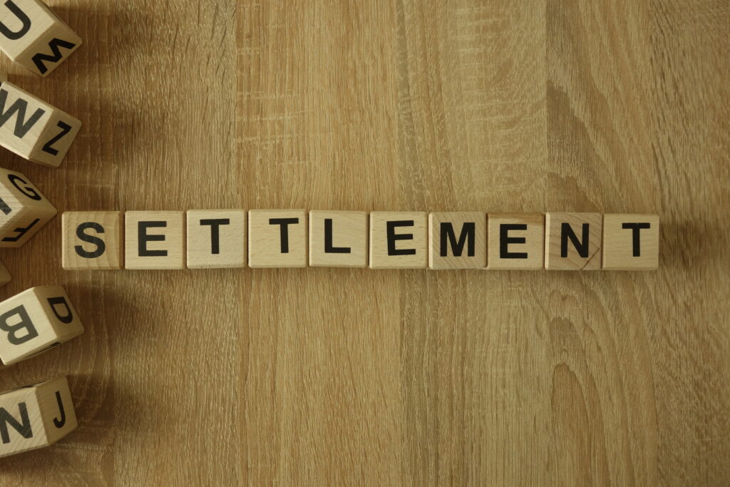 Wooden letter tiles spelling “SETTLEMENT” arranged on a wood surface.