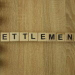 Wooden letter tiles spelling “SETTLEMENT” arranged on a wood surface.
