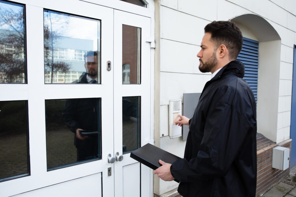 Person standing at a door and using an entry buzzer, carrying a document case.