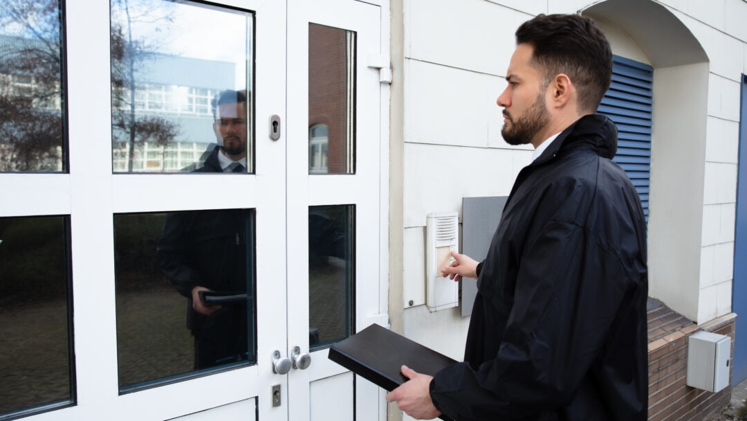 Person standing at a door and using an entry buzzer, carrying a document case.