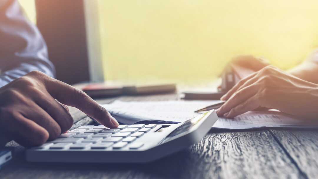 Close-up of hands using a calculator while reviewing paperwork on a desk.