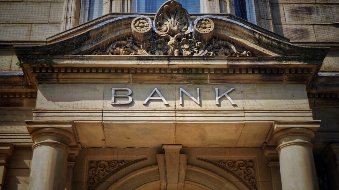 Stone bank building facade with columns and the word “BANK” displayed above the entrance.