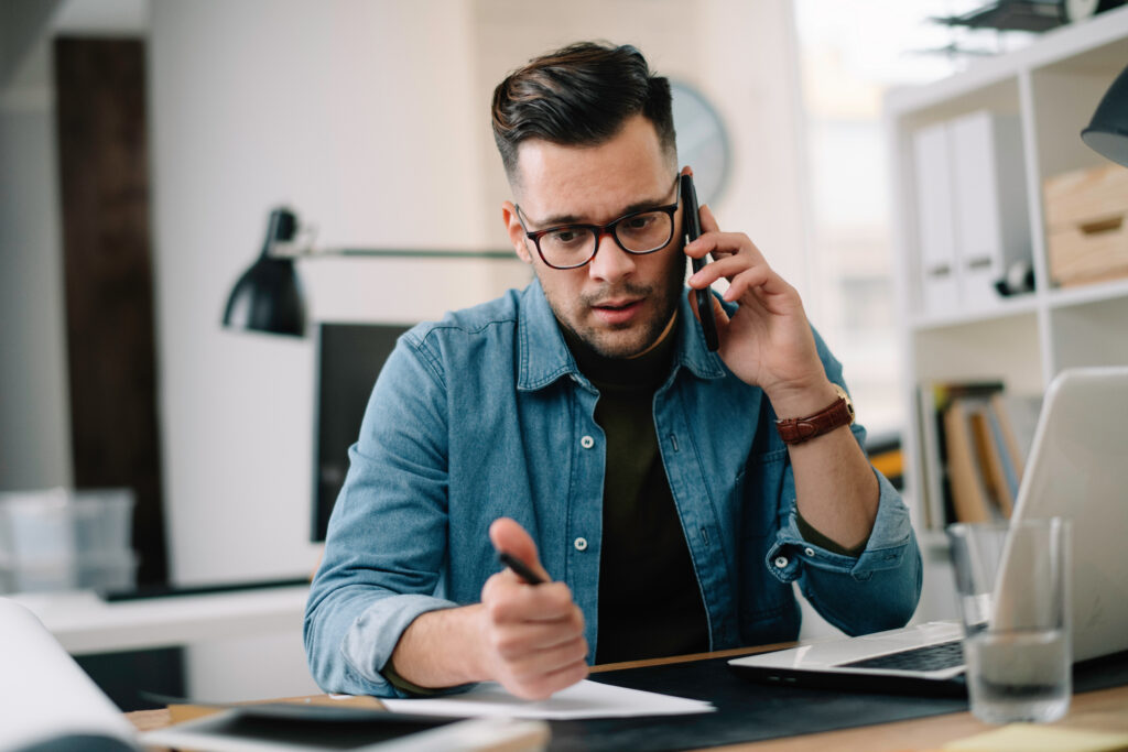 Man on the phone while reviewing paperwork and writing notes at a desk with a laptop.