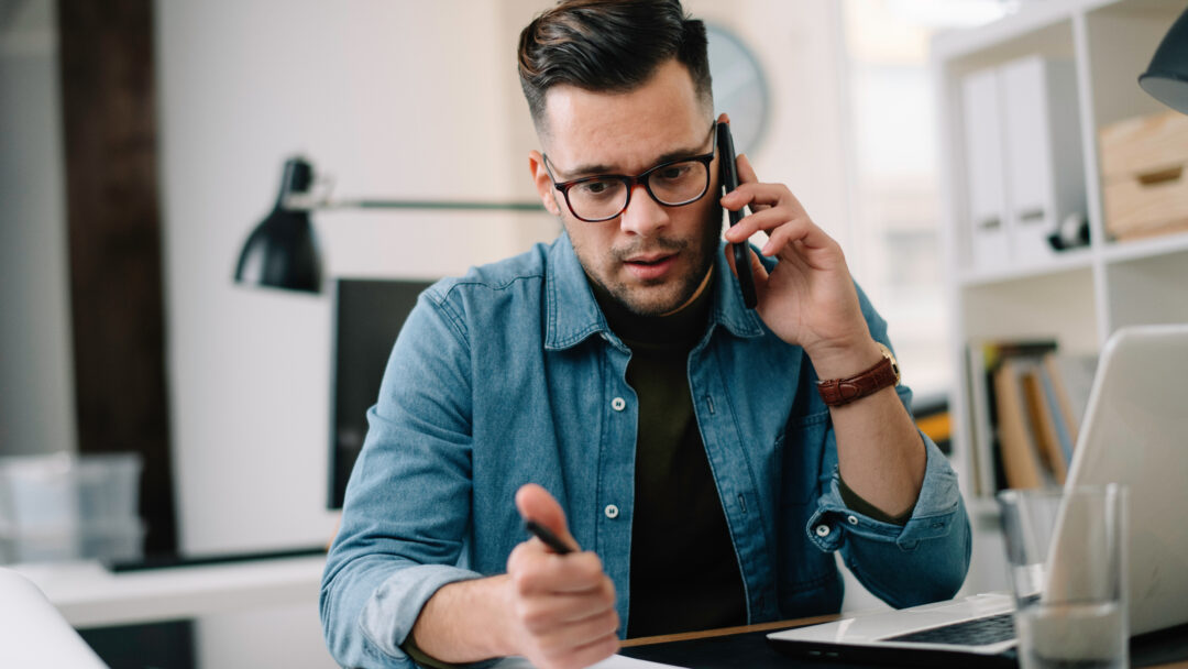 Man on the phone while reviewing paperwork and writing notes at a desk with a laptop.