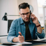 Man on the phone while reviewing paperwork and writing notes at a desk with a laptop.