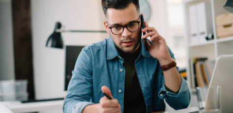 Man on the phone while reviewing paperwork and writing notes at a desk with a laptop.