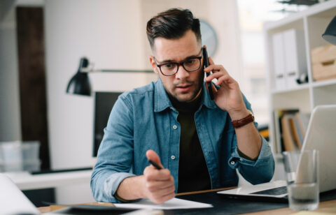 Man on the phone while reviewing paperwork and writing notes at a desk with a laptop.