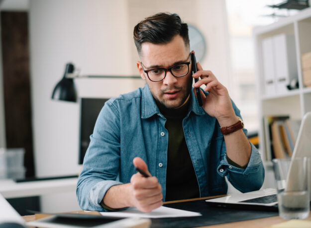 Man on the phone while reviewing paperwork and writing notes at a desk with a laptop.