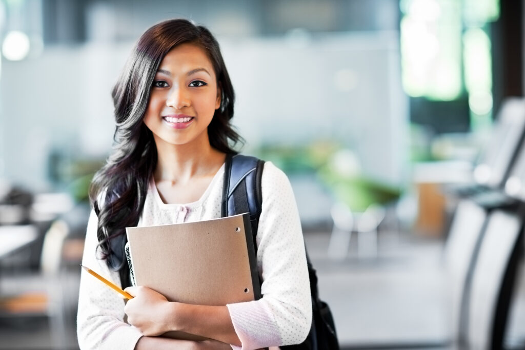 Young woman with a backpack holding a notebook and pencil, smiling in a school setting.
