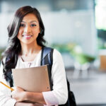 Young woman with a backpack holding a notebook and pencil, smiling in a school setting.
