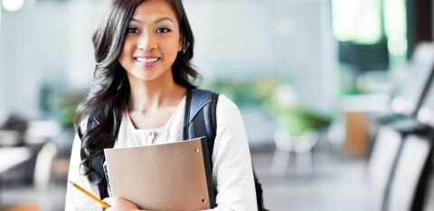 Young woman with a backpack holding a notebook and pencil, smiling in a school setting.