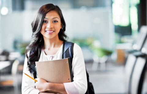 Young woman with a backpack holding a notebook and pencil, smiling in a school setting.