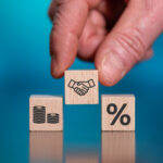 Hand placing a wooden block with a handshake icon between blocks showing money and a percentage symbol.