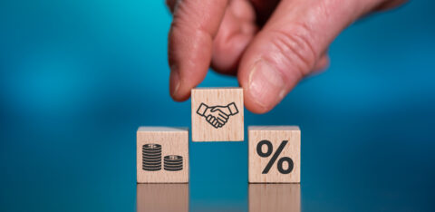 Hand placing a wooden block with a handshake icon between blocks showing money and a percentage symbol.