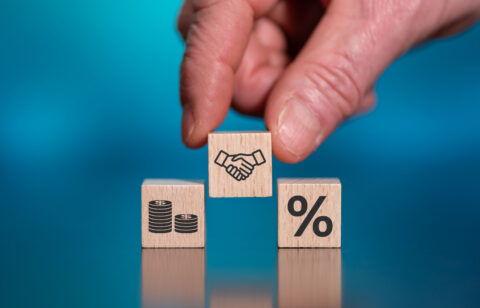 Hand placing a wooden block with a handshake icon between blocks showing money and a percentage symbol.