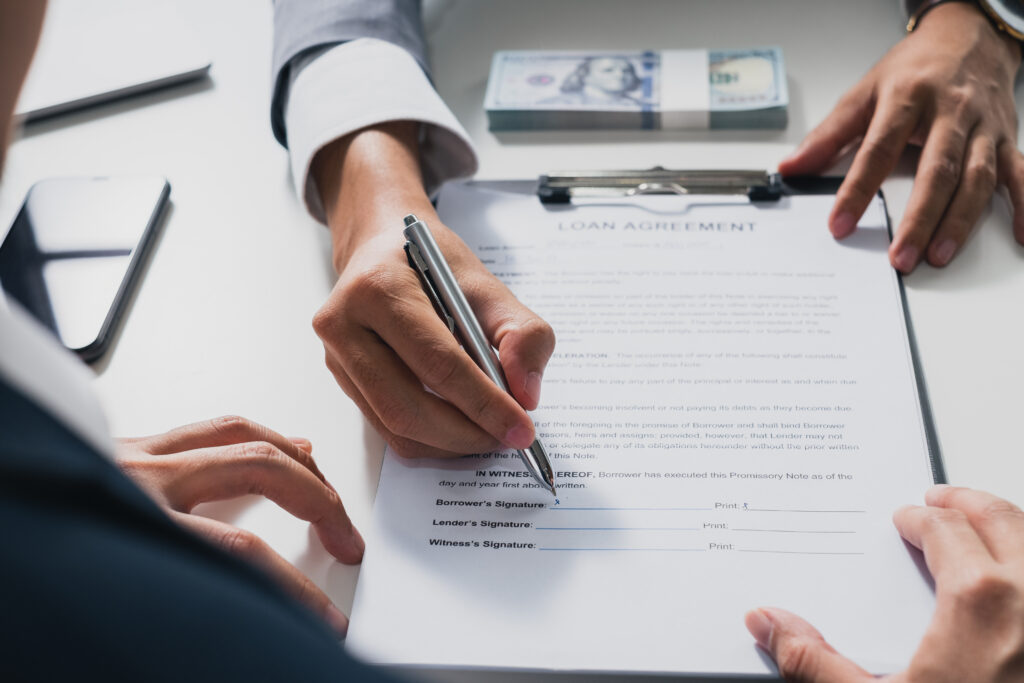 A person signs a loan agreement on a clipboard while another person points at the document, with a stack of cash nearby.