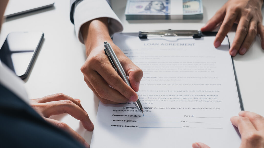 A person signs a loan agreement on a clipboard while another person points at the document, with a stack of cash nearby.
