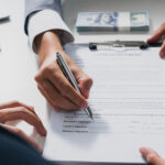 A person signs a loan agreement on a clipboard while another person points at the document, with a stack of cash nearby.