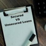 Clipboard with the words “Secured vs Unsecured Loans” next to a calculator and pen on a desk.