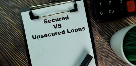 Clipboard with the words “Secured vs Unsecured Loans” next to a calculator and pen on a desk.