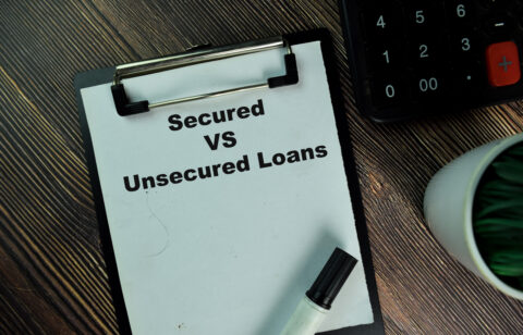 Clipboard with the words “Secured vs Unsecured Loans” next to a calculator and pen on a desk.