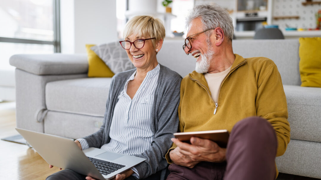 Older couple sitting on the floor at home, smiling while using a laptop and tablet.