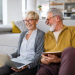 Older couple sitting on the floor at home, smiling while using a laptop and tablet.