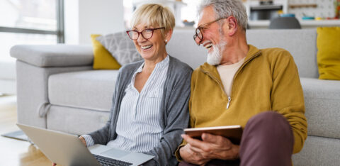 Older couple sitting on the floor at home, smiling while using a laptop and tablet.