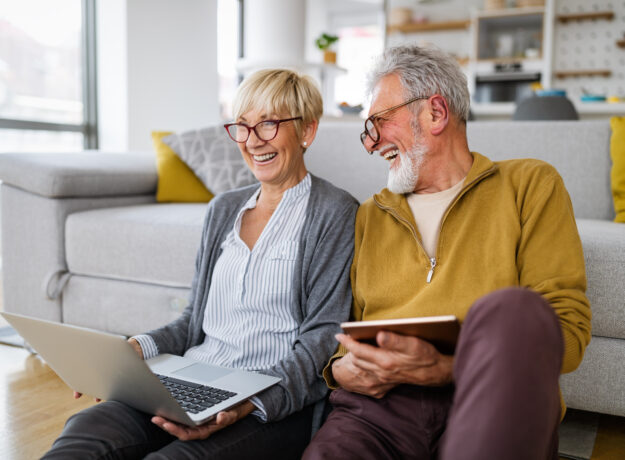 Older couple sitting on the floor at home, smiling while using a laptop and tablet.