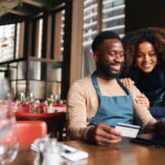 Pair of small business workers reviewing something online with a laptop and credit card inside a dining area.