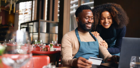 Pair of small business workers reviewing something online with a laptop and credit card inside a dining area.