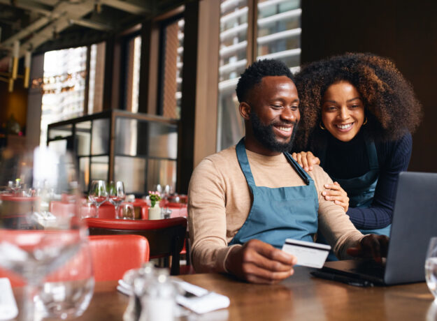 Pair of small business workers reviewing something online with a laptop and credit card inside a dining area.