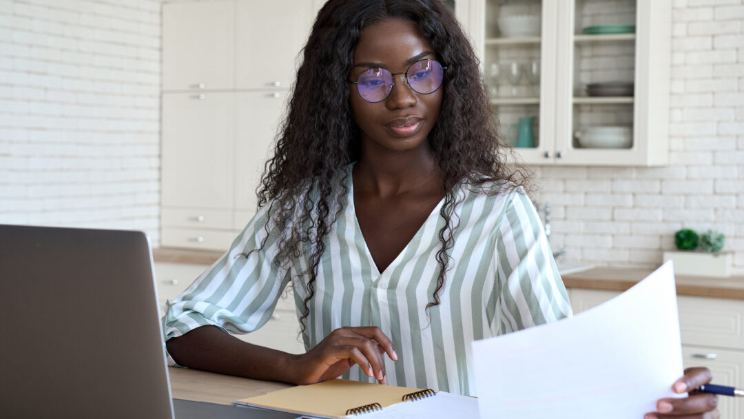 Woman reviewing a document at a table with a laptop and notebook nearby.