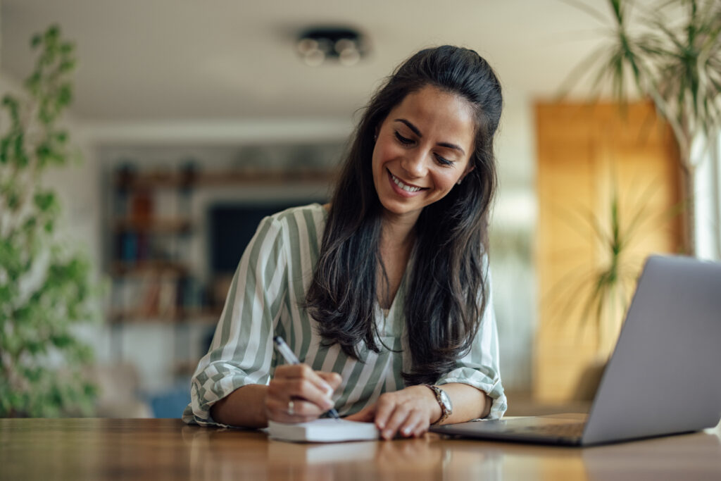 Woman smiling while writing in a notebook at a table with a laptop nearby.