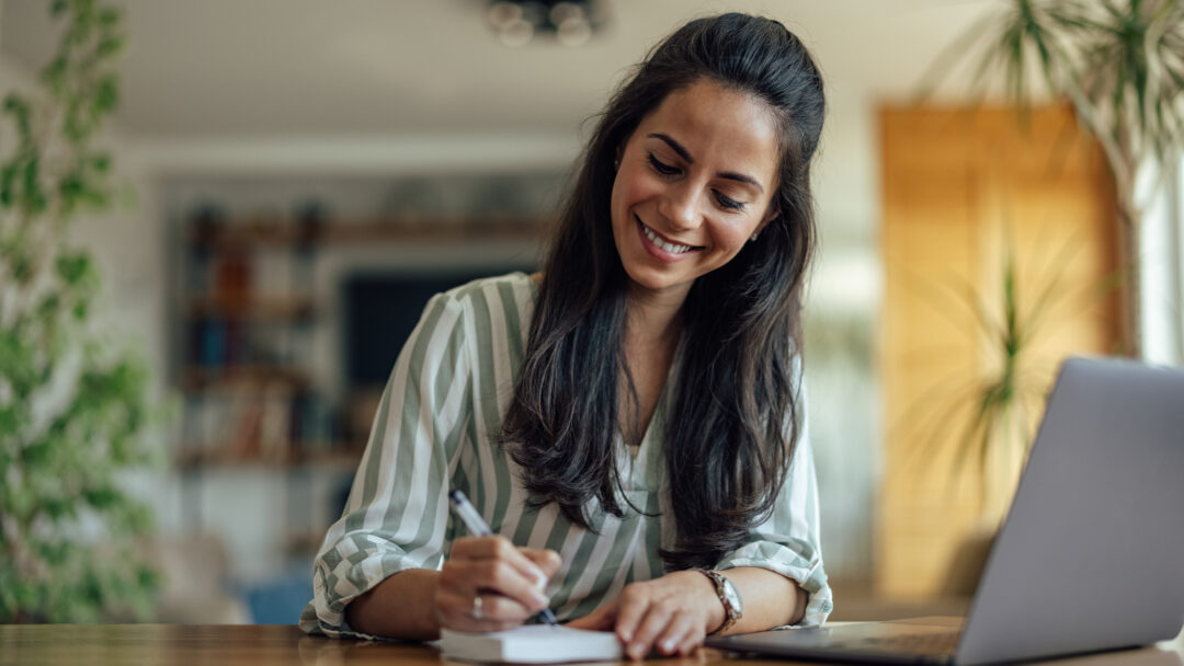 Woman smiling while writing in a notebook at a table with a laptop nearby.