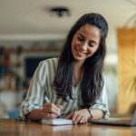Woman smiling while writing in a notebook at a table with a laptop nearby.