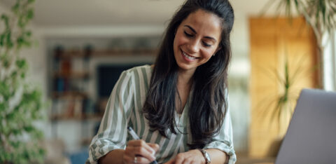 Woman smiling while writing in a notebook at a table with a laptop nearby.